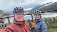 A woman (Margaux Dumas) and a man (Jonas Pollard) both wearing cycling helmets and waterproof jackets. The woman's jacket is blue and the man's is red. In the background is a large body of water and a mountain. 
