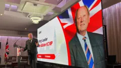 Jim Allister is wearing a suit and talking on a platform. He's looking to his side at a large screen. The screen shows an image of Allister with the union flag behind him.