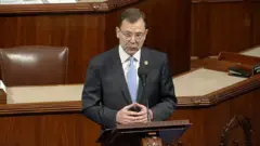 A man in a suit stands at a podium holding his hands together