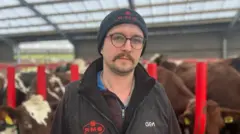 Farmer Adam Johnstone, wearing black wool, gilet and hat, stood with a sad face in front of a herd of cows in a dairy shed