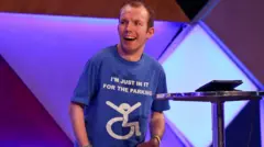 Tricia Yourkevich/BBC Lee Ridley smiling and standing on a comedy stage. He has cerebral palsy and uses a text-to-voice synthesiser. He wears a blue shirt featuring a drawing of someone in a wheelchair, captioned: "I'm just in it for the parking".