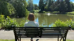 A woman is sat on a bench next to a lake. You can see the back of her body as she looks at the water. There are green plants around the water's edge, trees in the background and a fountain in the middle of the lake.
