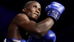 Nuhu Batte, with blue boxing gloves on and with an arm flexed, preparing to fight.