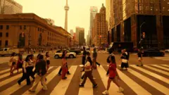 People walk on a crossing in a road downtown Toronto. There are buildings in the background and sky is orange from smoke.