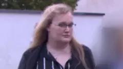 Rose Henderson arrives at the tribunal. Rose has long blonde hair, glasses and wears a black and white striped dress. What looks like a black jacket or cardigan over that but the image is blurry so it's not clear.