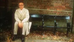 Alfred Brendel sitting on a park bench, wearing a trench coat