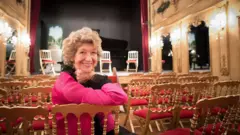 Dame Felicity Lott with short curly hair dressed in black and pink leans on the back of a chair and smiles to a camera while sitting in a theatre space.