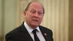 Liam McBurney/PA TUV leader Jim Allister is an older man with fair hair. He is wearing a dark jacket and tie which is black with white spots and a white shirt. There is a badge on his left lapel which is red with white lettering. He is standing in front of a beige background.