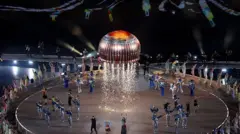 A giant teacake, wrapped in red and silver packaging, rises into the sky with sparks firing from underneath it. Various dancers and performers stand beneath it on the floor, performing a routine. 