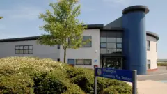 The two storey modern building is made of various blue, white and brown materials. There is a tall blue column running up the middle. The building is set in from the main road, behind a pavement area with hedges and a tree on it. A sign in the foreground reads: "Washington Community Fire Station."