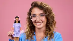 Mattel/PA A woman with wavy light brown/blonde hair smiles while holding a Barbie doll with brown hair and pink ear protectors. She is wearing glasses and a light blue top and is posing in front of a plain pink background.