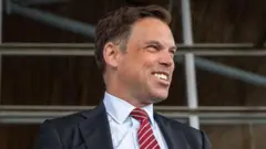 Ken Skates smiling off to the side of the image, wearing a dark suit and red and white tie, with a pin stripe shirt.