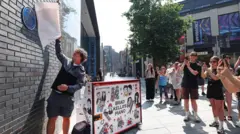 Brad Kella is standing, and pulls a white cover from a blue plaque as people watch and take photographs on their phones beside a decorated piano. It is sunny day. 