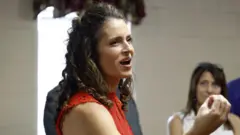 Julia Letlow is seen talking to constituents which smiling and appearing to talk. She is turned to the side and is seen from the shoulder up. She is wearing a red top.