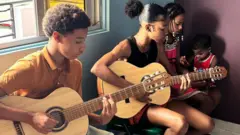 Cape Verdean children sit down and play guitars. Three are sat in a line.