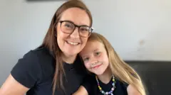 Mum Sara is smiling and wearing glasses. She is alongside her young daughter Ellie who is smiling and wearing a multi-coloured necklace. They are pictured at home when Ellie is educated by her mum.