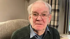 Alan is looking in to the camera. He wears glasses. He has a jumper on with a checked shirt underneath. He's sitting on a beige sofa in a living room. A tall lamp is behind him. The curtains are checked.