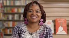 A woman with reddish-brown hair and pink comb-shaped earrings smiles at the camera, wearing a pink and blue checked top with white embroidered trim. She's in a bookshop, with blurred shelves of books on the left and a display featuring copies her books on the right.