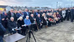 People are sitting and a crowd is standing. A man is holding a wreath. Some are holding roses.