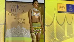 The contestant of the beauty contest in Daloa in the mad bikini and a short shirt with traditional designs and holding the fly-wave flying. Behind it are banners in yellow with Miss Cote D'Ivoire 2025 logo.