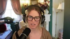 BBC A woman, QTCinderella, is sitting wearing black over-ear headphones and black glasses, with a brown, open hoodie. She is speaking into a black microphone. In the background is a lamp and a vase of flowers and a window with pink curtains.