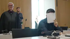 A man holds a sheet of paper over his face, wearing a black shirt in court