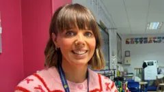 BBC Stacey smiling at the camera. She is wearing gold hoop earrings and a pink and red patterned cardigan. She has short brown hair and a fringe.