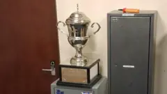 A large silver cup, with etchings of greyhound-type dogs and hares, on a wooden base with silver plating around it. The cup stands on some utilitarian-looking cabinets which are used for storing ammunition, in a non-descript office.