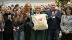 Reuters Britain's Queen Camilla holds a tote bag with the name of Jilly Cooper on it, who wrote the original books entitled Rivals, as she poses with members of the cast and crew during a visit to the set of the TV series Rivals.