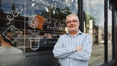 Willi's Cafe owner Rod Davis stands in front of a window covered in writing and doodles, including a drawing of a plant pot. He is wearing glasses and a blue/grey striped shirt. There are customers inside the cafe and street reflections in the glass.