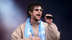 Tom Grennan is wearing dark glasses and singing into a microphone. He is wearing a light beige jacket and Coventry City scarf around his neck