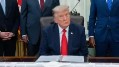 US President Donald Trump sitting in the White House wearing a blue suit jacket, white shirt and red tie.