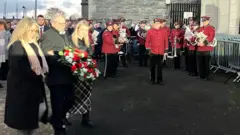 A man is holding a wreath. He's being supported by two women. A band are wearing maroon uniforms and holding instruments. A crowd of people are watching.