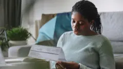 A woman sat on a living room floor looking at a heating bill. She is wearing a light green jumper and behind her is a grey sofa, a plant on a table, and a white wall.