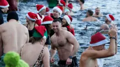 The PA media image shows swimmers in their bathing suits and Christmas hats taking part in the Christmas Day dip at Blackroot Pool, Sutton Park in Sutton Coldfield, Birmingham on December 25, 2025.