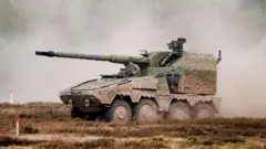 A large military vehicle with four wheels on one side. It is covered in brown and green camo paint. On top, there is a long thin gun barrel. There is smoke all around it