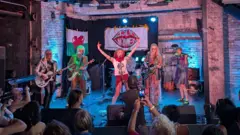 Six women dressed in punk clothing stood on a stage playing instruments. In front of them is a crows, many of whom are holding up cameras and phones. Behind them are two flags hanging on a wall, one is a Welsh flag and the other is a white flag with a large red lips logo which say "Loud Women".