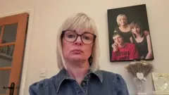 Sharon Huddleston, with blonde hair and glasses, looks sadly into the camera. On the wall behind him is a photo of him with his two children.