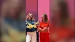 Nuala McGovern and Katie Weir stand side by side each holding a bra. They both smile at the camera.