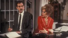 John Cleese & Prunella Scales in Fawlty Towers. They are shown arguing in the hotel lobby behind the front desk.
