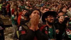 A man is holding his hands over his mouth with numerous of Mexican fans behind him.