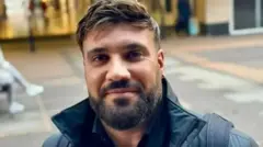 Close up head and shoulders shot of Alastair Chambers wearing a black coat. He has a beard and a moustache. He is standing in a pedestrianised shopping area.