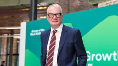 West Midlands Mayor Richard Parker, wearing a navy blue suit and a white shirt with a red and white striped tie. He is standing in front of a green and blue branded board.