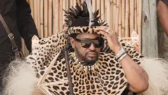 King Misizulu wearing traditional leopard-skin clothing and holding a stick. He is wearing dark glasses and is touching his head