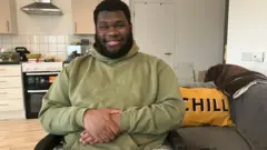 A young man with dark hair and beard wearing a green hooded sweatshirt sits in front of the camera