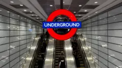 BBC A large Tube roundel hangs above a bank of three escalators as Euston underground station. There are white tiles on the wall and people are visible riding the escalators.