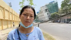 The lady stands outside, looking straight on the camera, and in the background is a white building, dangerously leaning toward one side