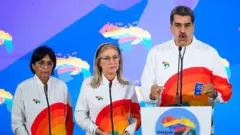 Gaby Ora/Bloomberg via Getty Images Nicolas Maduro, right, speaks to members of the media next to Cilia Flores, center, and Delsey Rodriguez after voting during a referendum vote in Caracas, Venezuela, on Sunday, December 3, 2023. He is wearing a matching track suit with rainbow printed on it.
