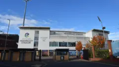 The exterior of Headingley Stadium is shown with its large white panels, glass windows and tall floodlights rising above the building. A gated entrance and autumn‑coloured trees sit in the foreground, making the stadium look both modern and familiar as a major Leeds sports venue.