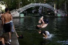 A topless young man with brown hair wearing black shorts and trainers somersaults into the water at Canal St Martin.